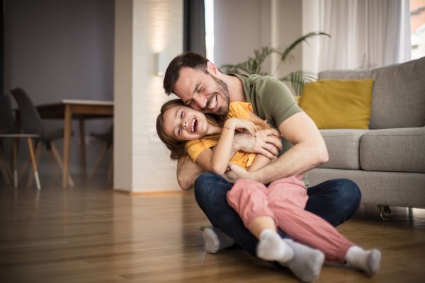 A father hugging his laughing young daughter while sitting on an apartment floor